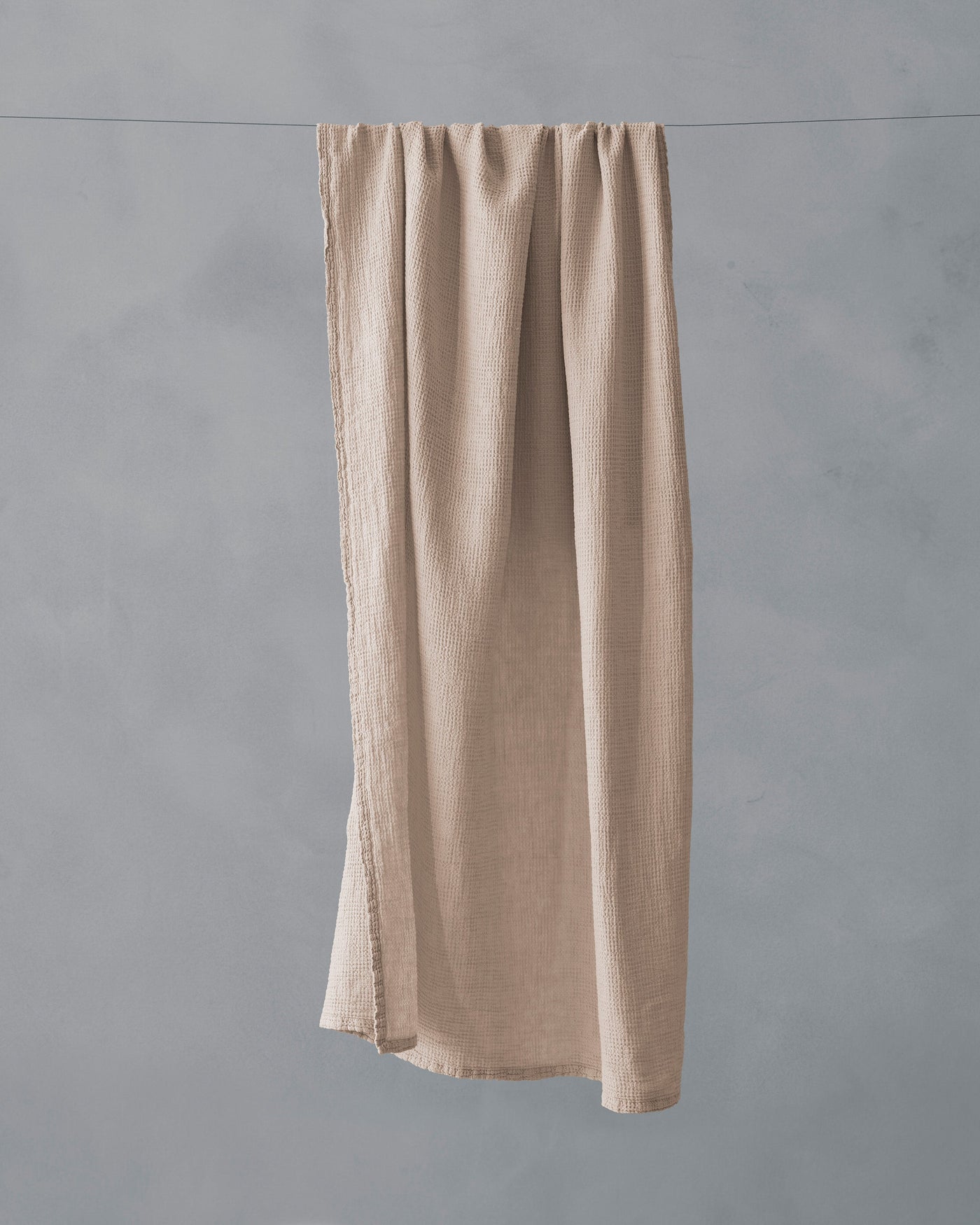 Sand-toned linen bath towel hanging full-length on a line, showing natural texture and frayed edges.