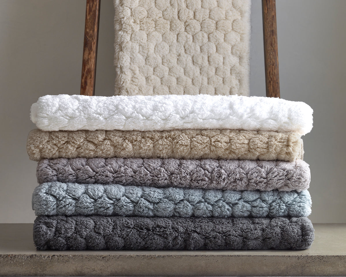 Matouk Lotus bath rugs stacked in ivory, white, taupe, lavender, sky, and slate.