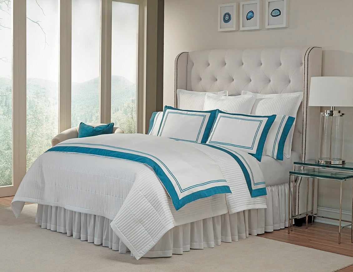 White and teal embroidered border bedding styled on a tufted bed in a bright bedroom.