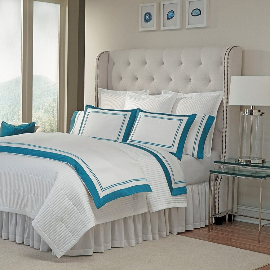 Home Treasures Marco white bedding with teal border on bed with tufted headboard.