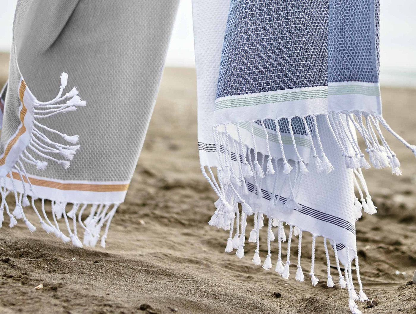 Close-up of Coyuchi Mediterranean towel fringe and stripe detail on sandy beach.