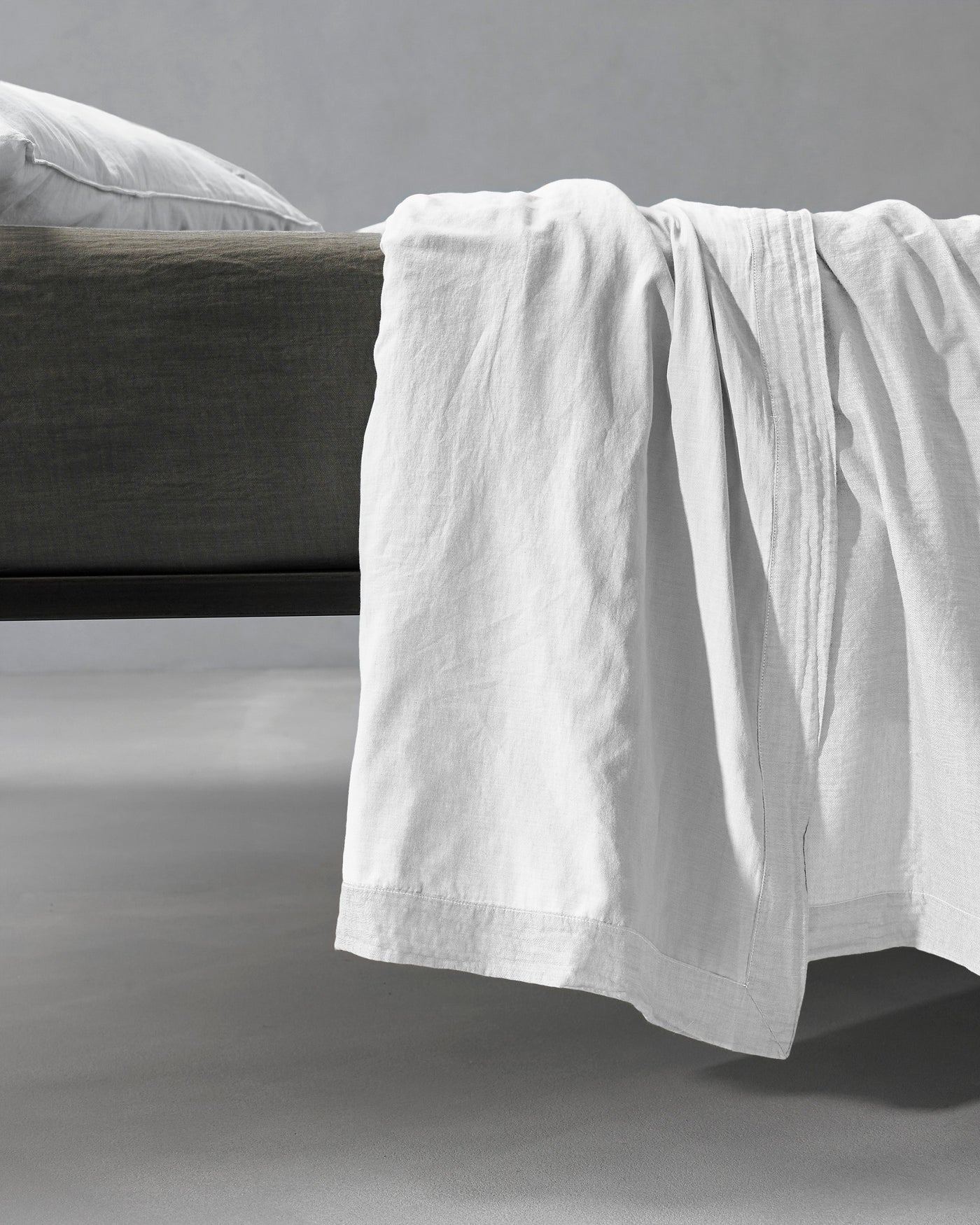 Society Limonta Miro plain flat sheet in white linen draped over the edge of a dark bed frame.