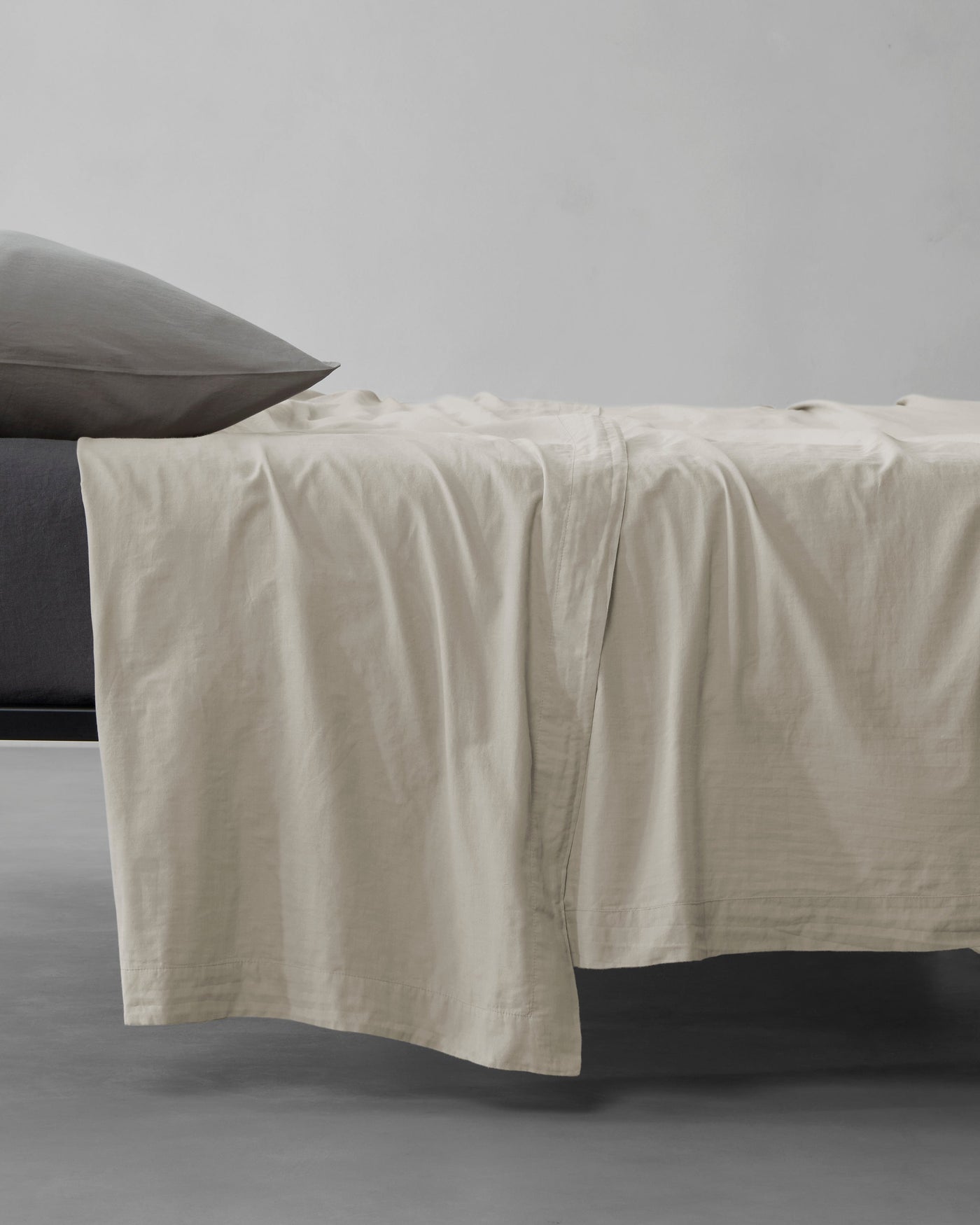 Society Limonta Miro flat sheet in sand linen, draped over bed edge with charcoal pillowcase.