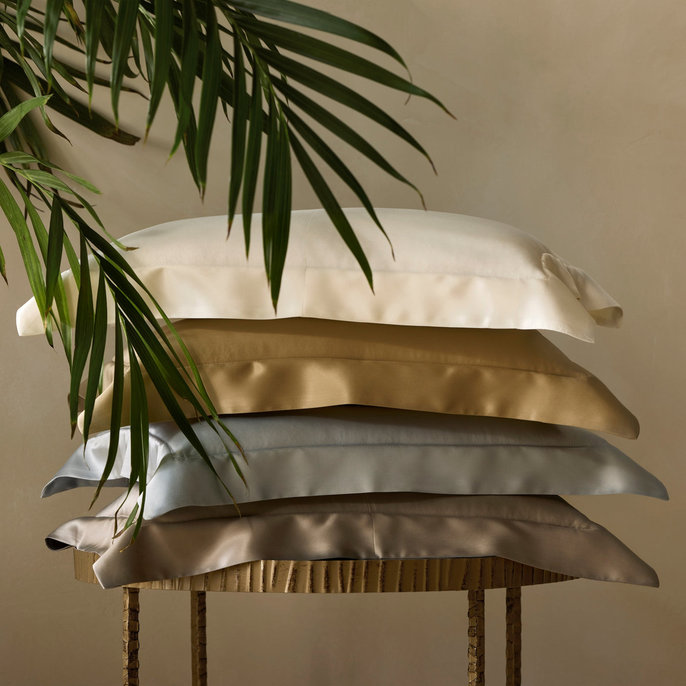 Four stacked silk pillowcases in ivory, champagne, silver, and taupe on a gilded stand.