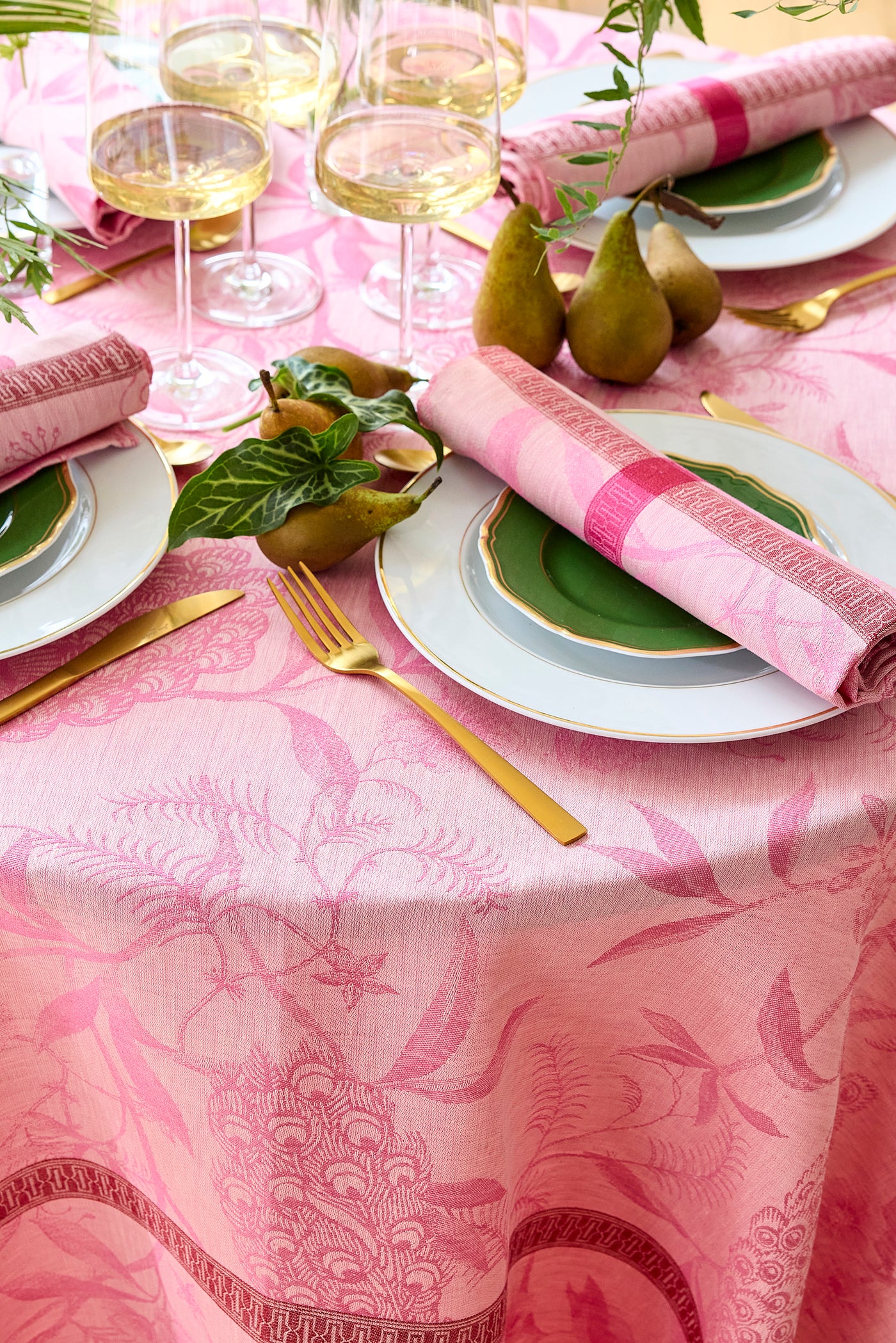 Close-up of pink jacquard tablecloth with botanical motif, styled with gold cutlery and green plates.