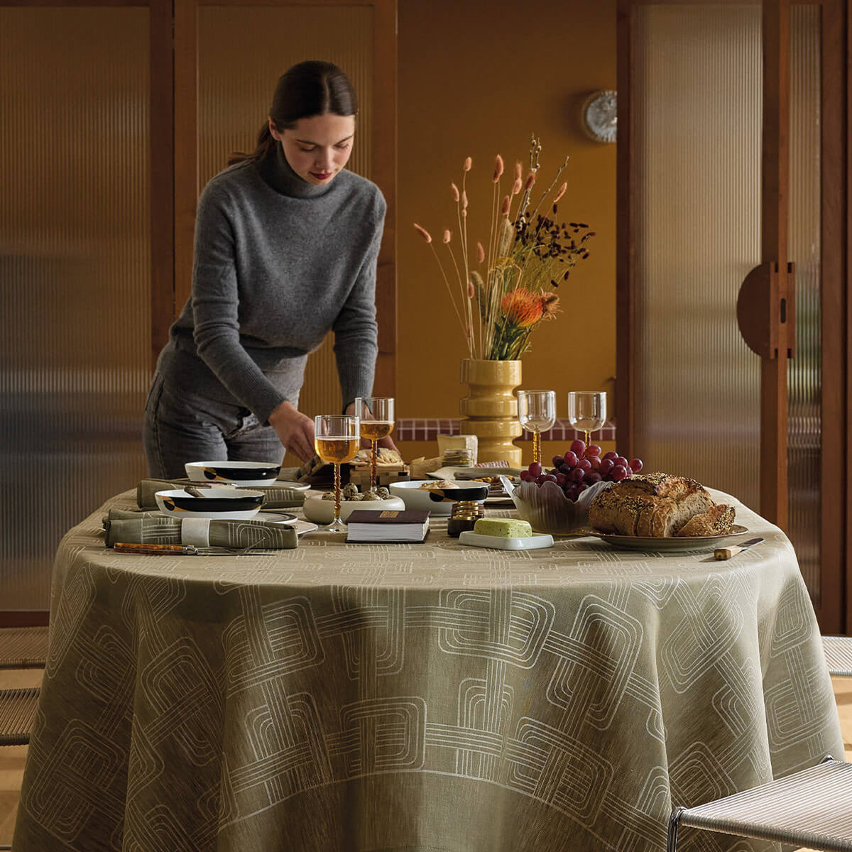 Woman arranging a styled table set with Le Jacquard Français green jacquard tablecloth, food and glassware.