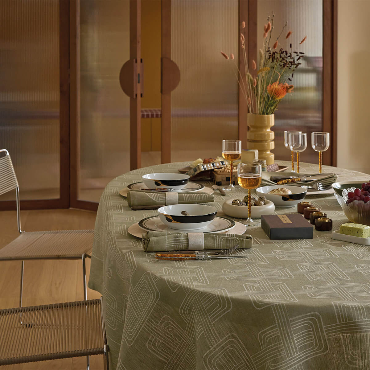 Oval dining table draped in Le Jacquard Français Green jacquard tablecloth with geometric motif, set for dinner.