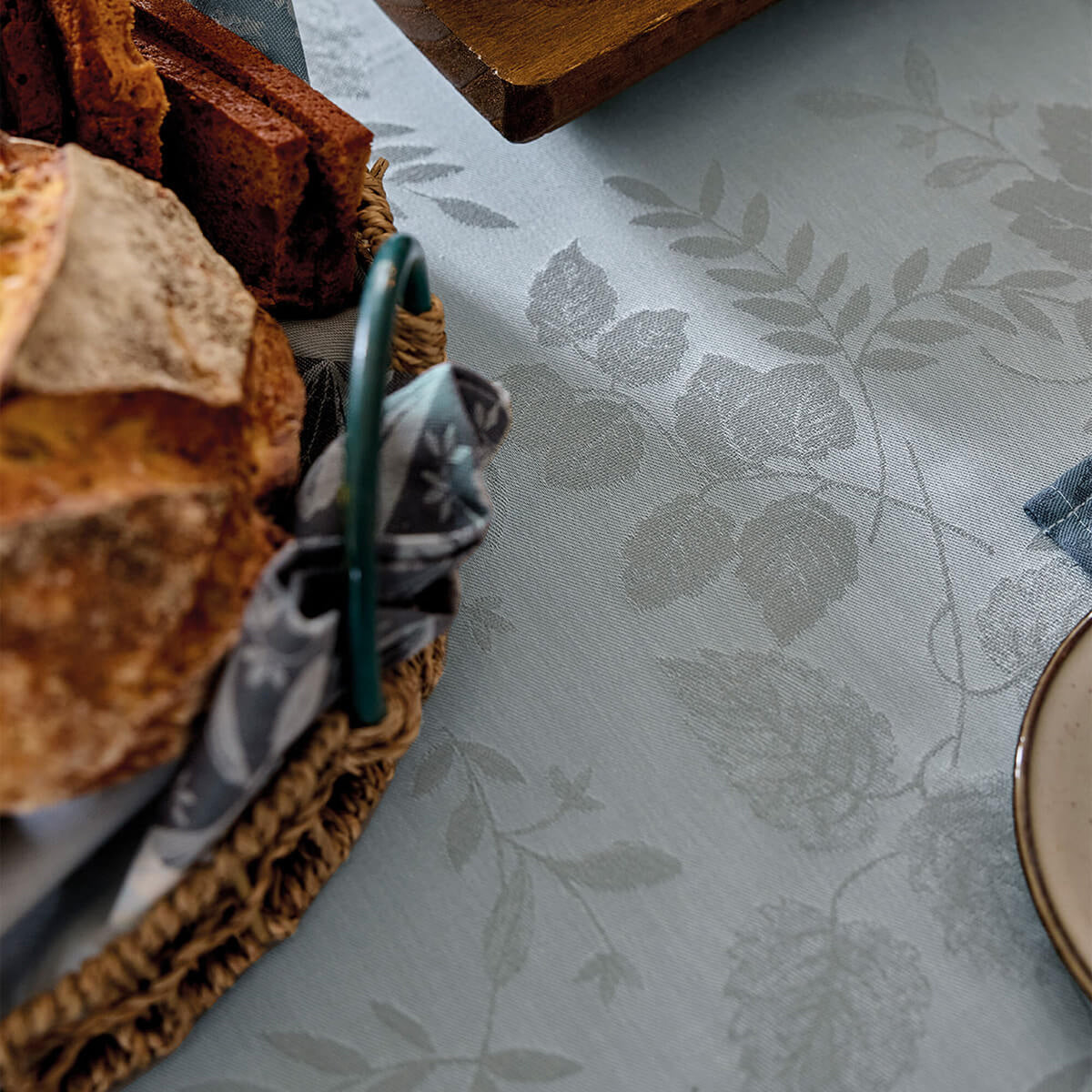 Close-up of Le Jacquard Français Manor Lane tablecloth in Blue, showing woven floral jacquard pattern styled with bread basket.