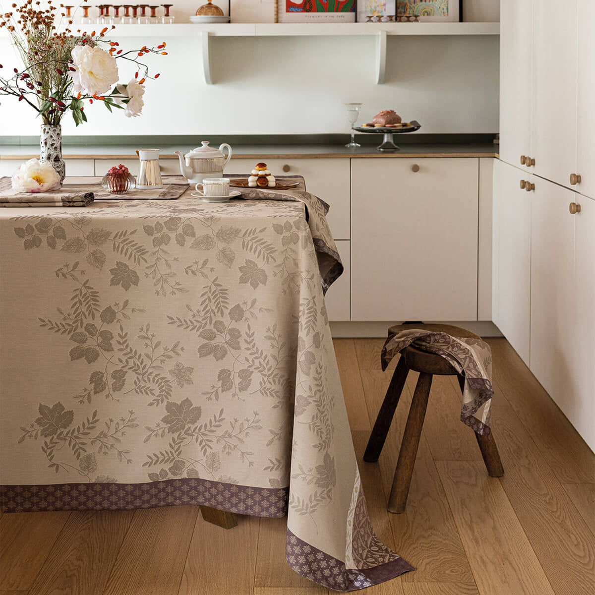 Beige tablecloth with tonal leaf jacquard and floral border, styled on a dining table in a modern kitchen.