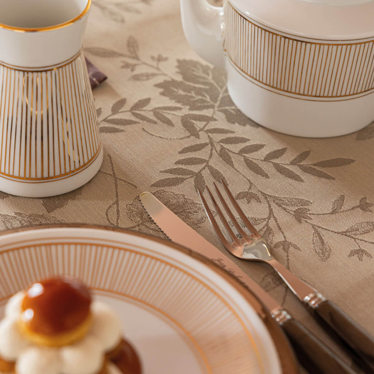 Beige tablecloth with woven leaf motif, styled with striped gold-rimmed china and silverware.
