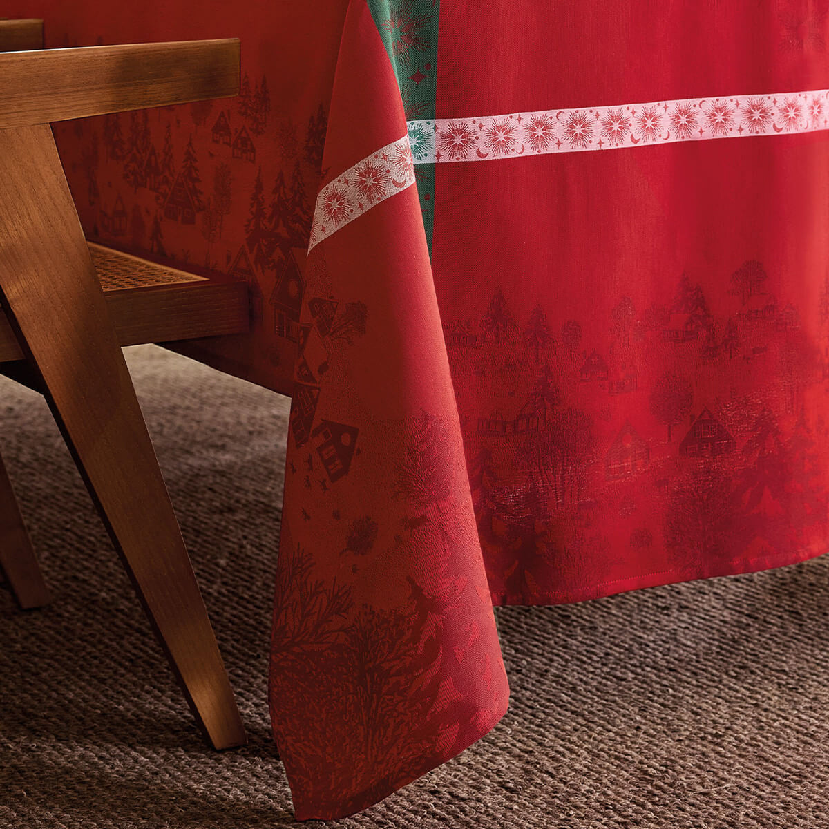 Red tablecloth corner draped over wooden table, toile holiday motifs and snowflake border detail.