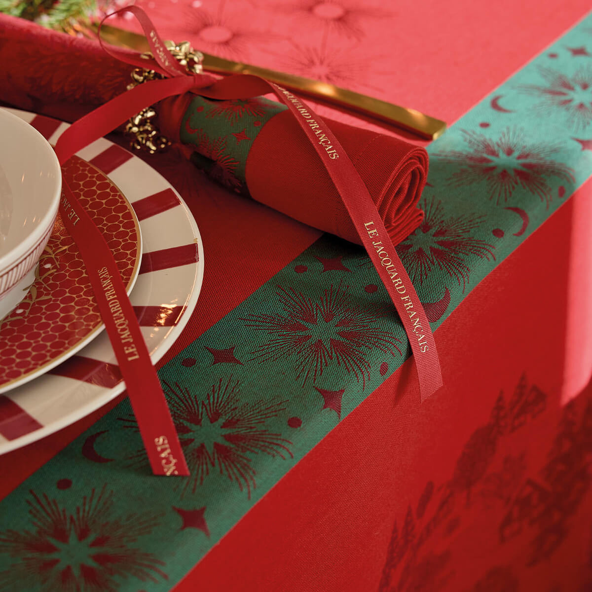Red and green jacquard tablecloth with gold star pattern, styled with holiday place setting and ribbon.