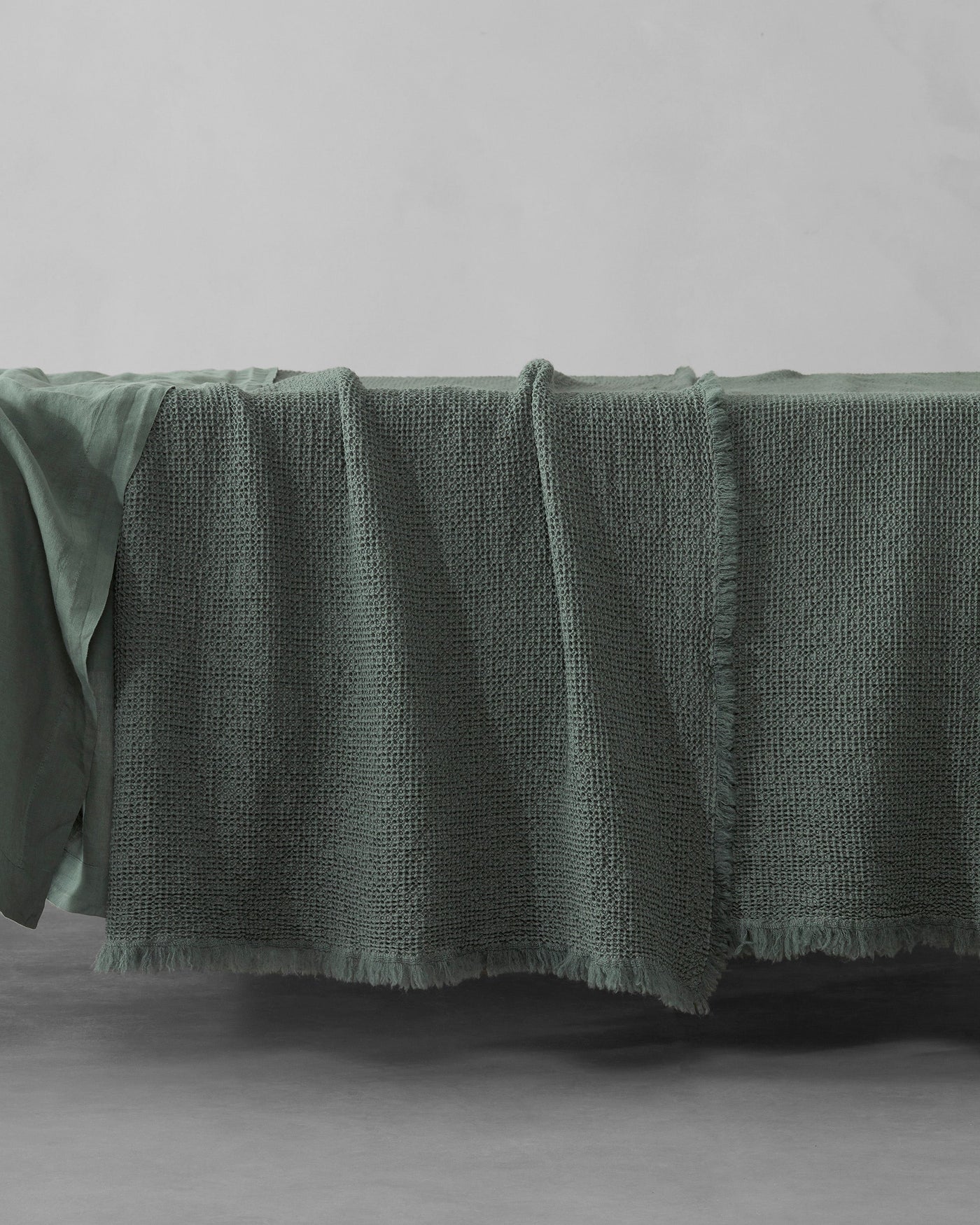 Sage green textured waffle-weave blanket with fringe edge, draped over a bed.