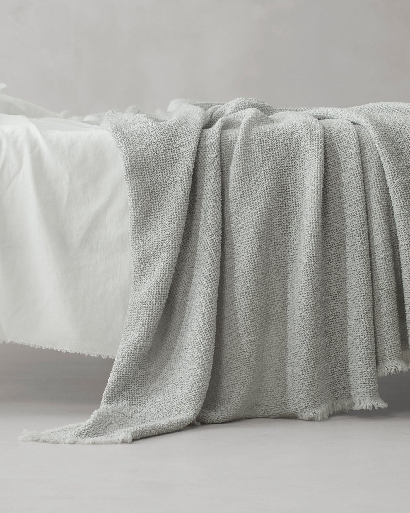 Textured woven blanket in mist grey draped over bed edge, fringe hem detail visible.