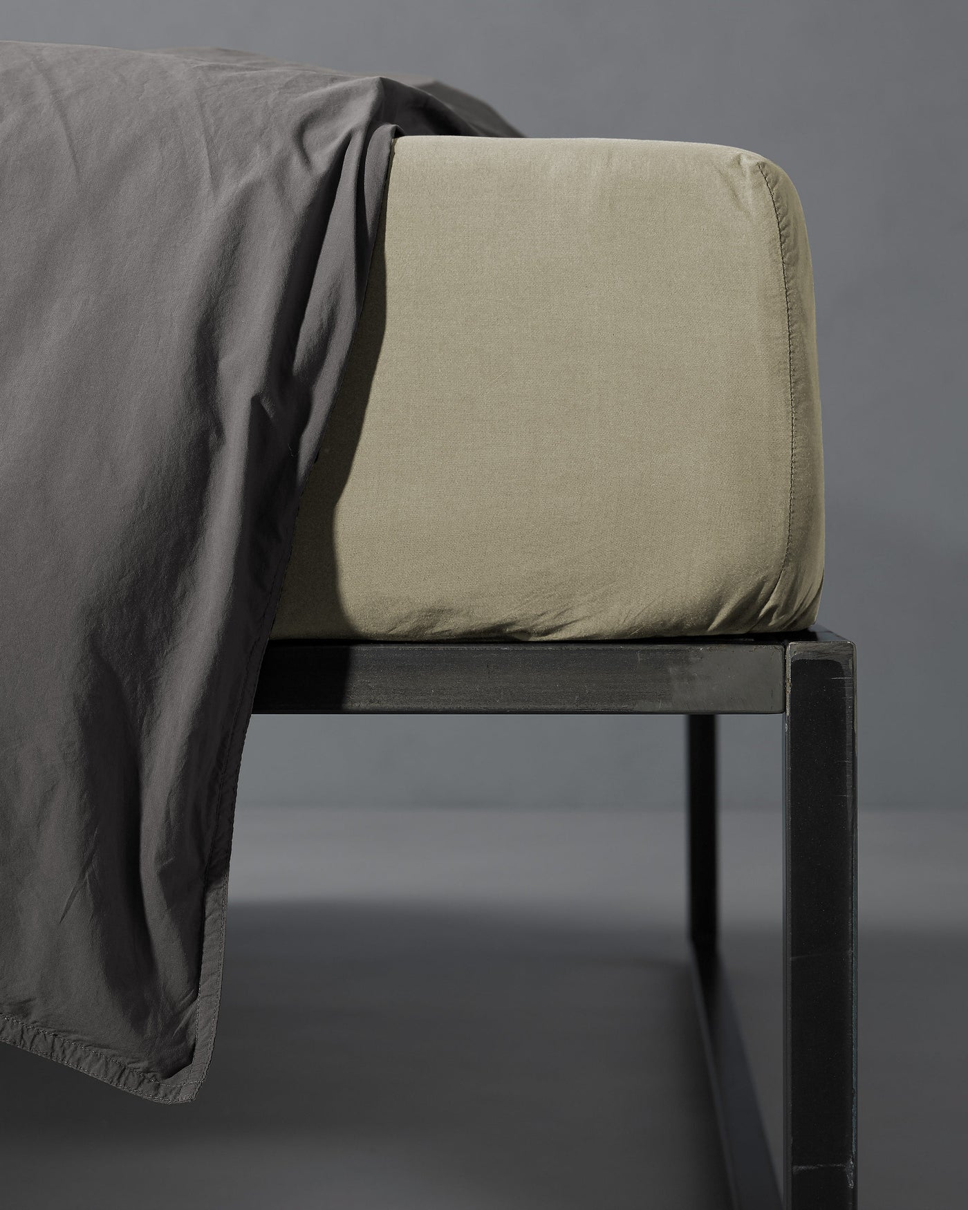 Corner detail of sage fitted sheet on mattress with charcoal flat sheet draped over black bed frame.