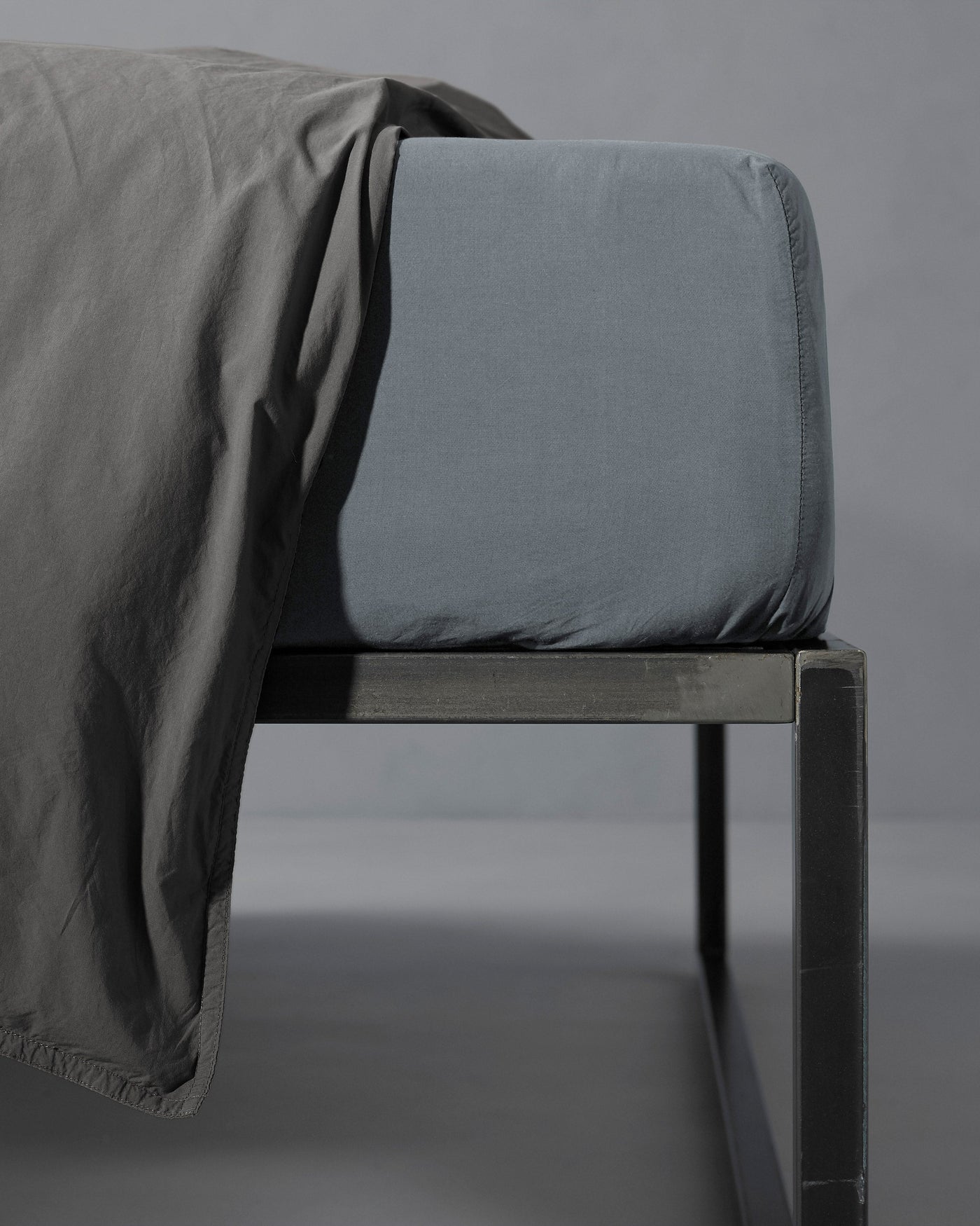 Close-up of slate blue fitted sheet corner on metal bed frame, charcoal flat sheet draped over.