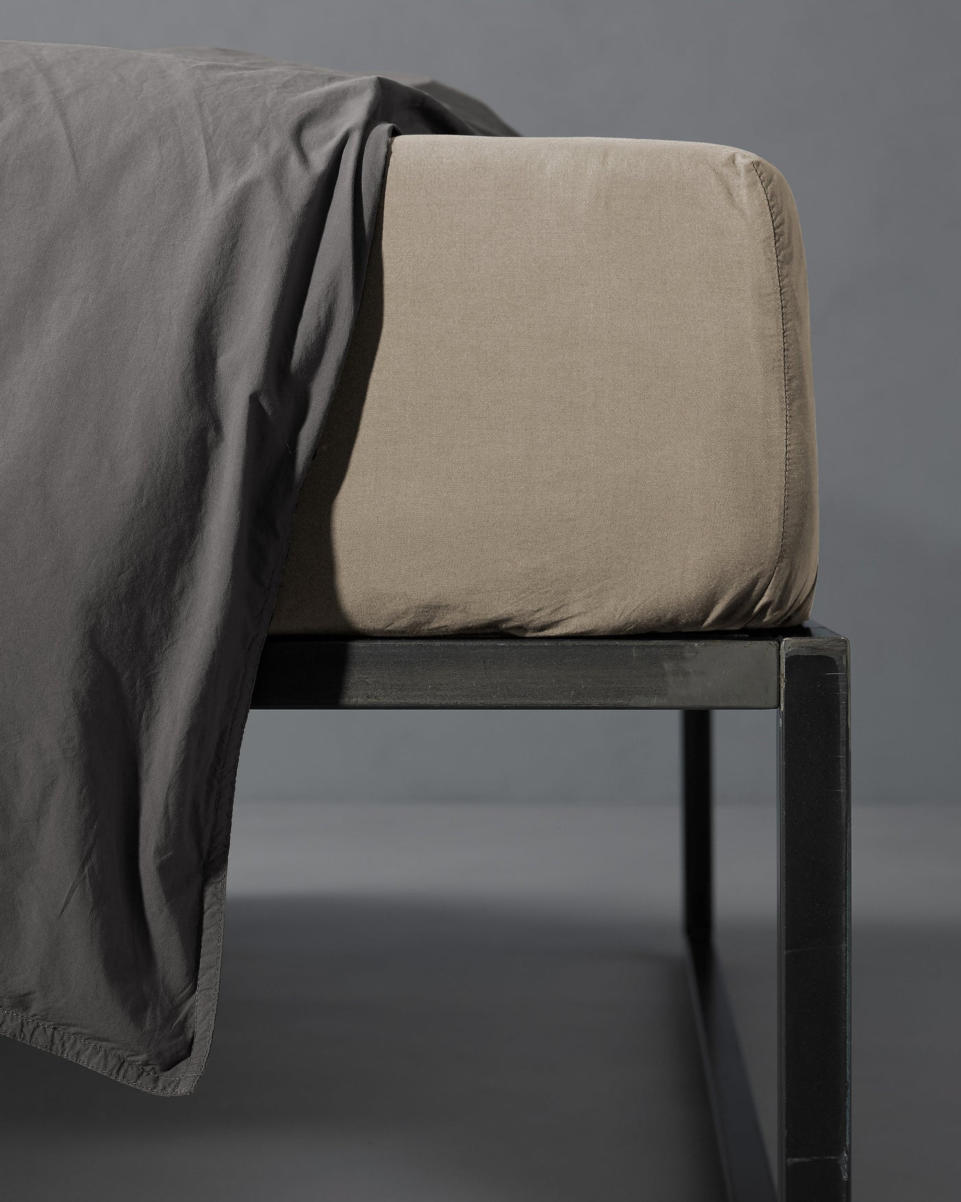 Close-up of sand fitted sheet corners on mattress with charcoal flat sheet draped over bed frame.