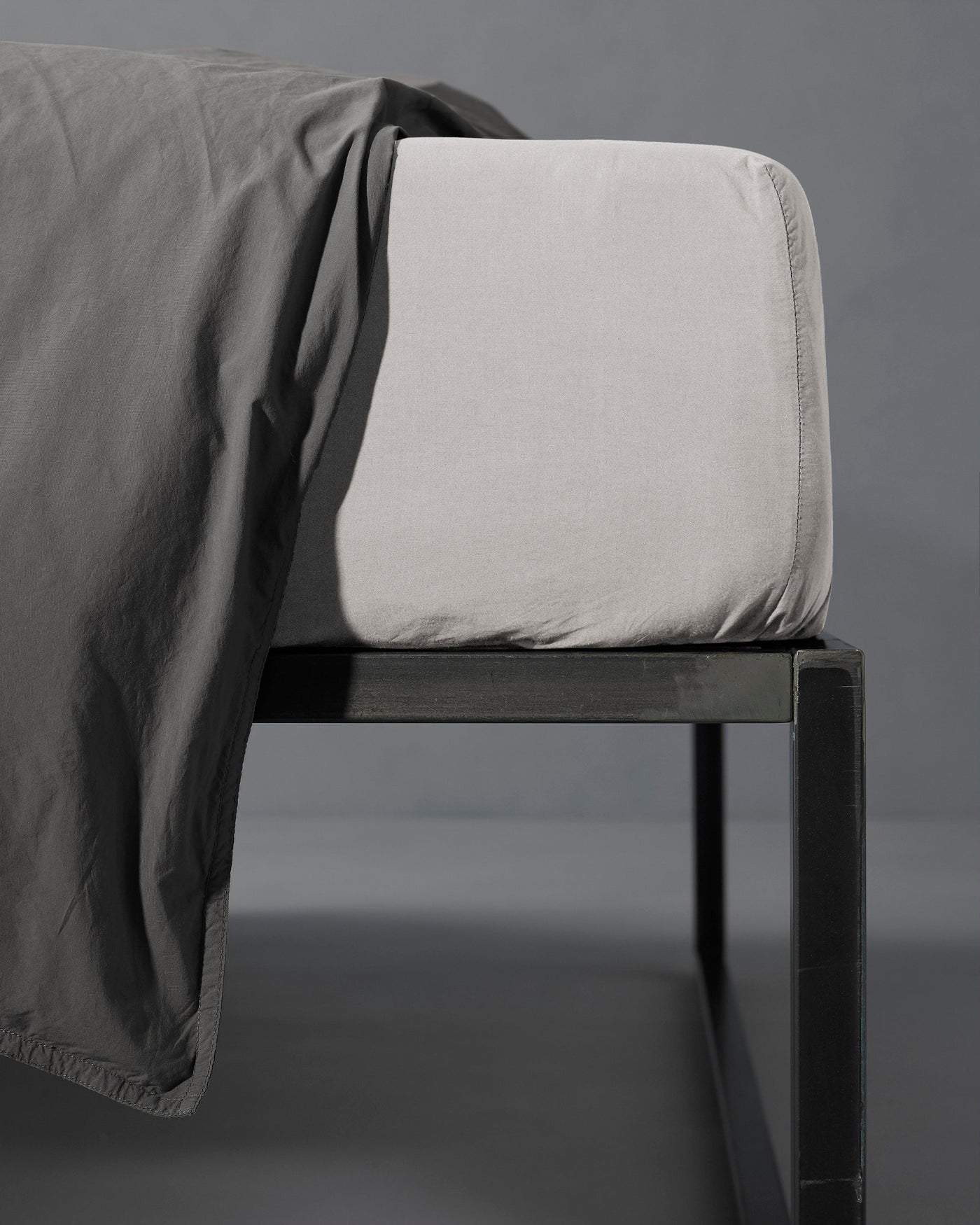 Corner detail of a pebble fitted sheet on a dark metal bed frame, with slate flat sheet draped over.