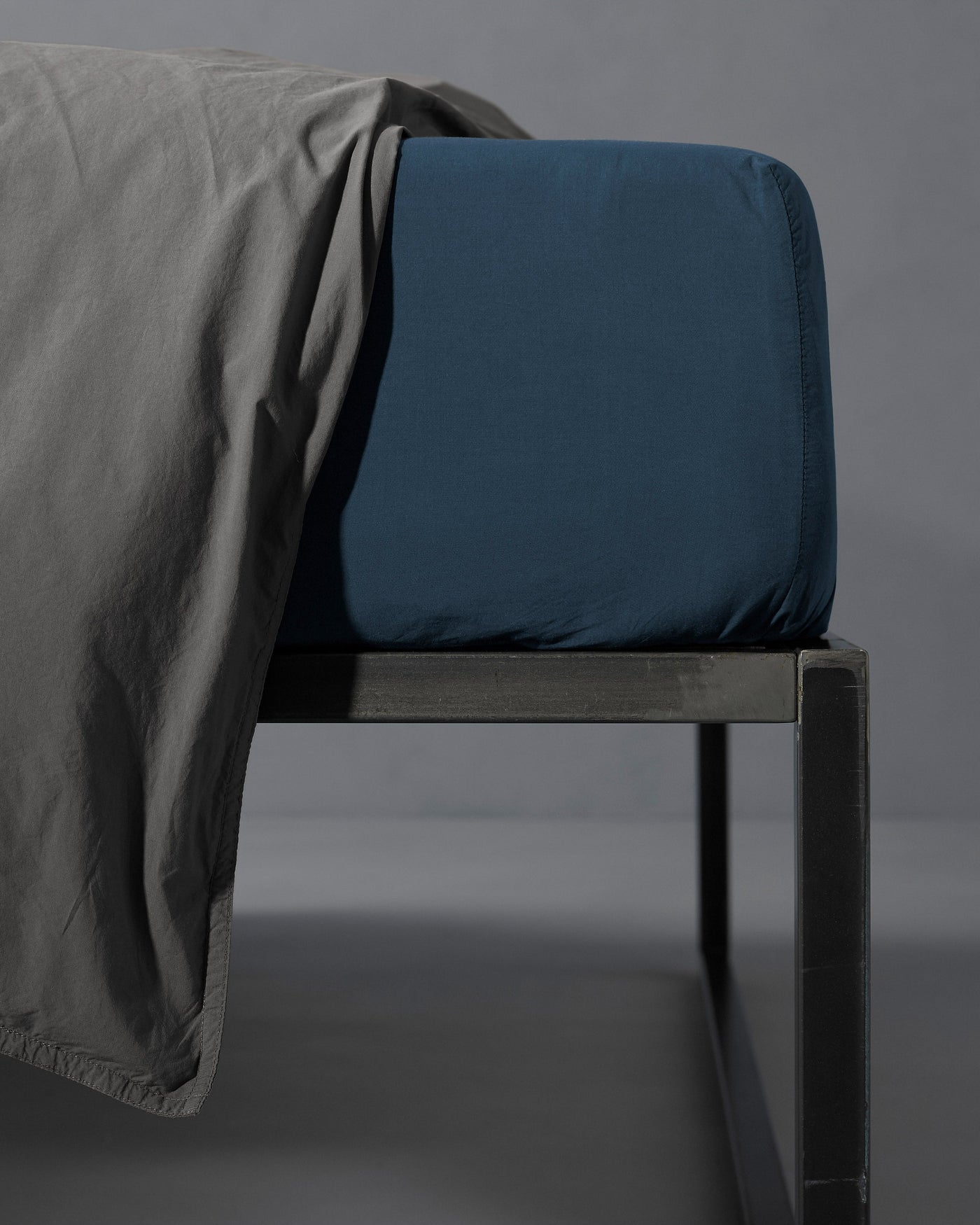 Society Limonta Nite fitted sheet in deep teal on bed frame with grey draped sheet.