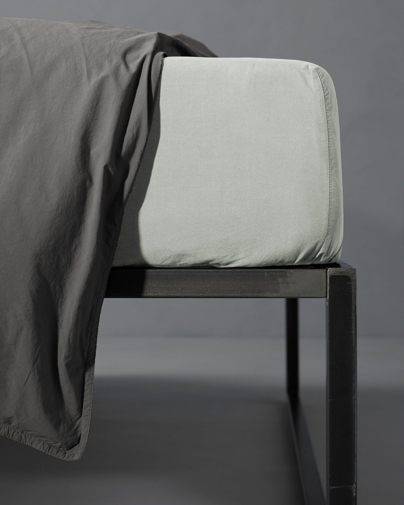 Corner detail of a sage fitted sheet on a mattress with a charcoal flat sheet draped over.