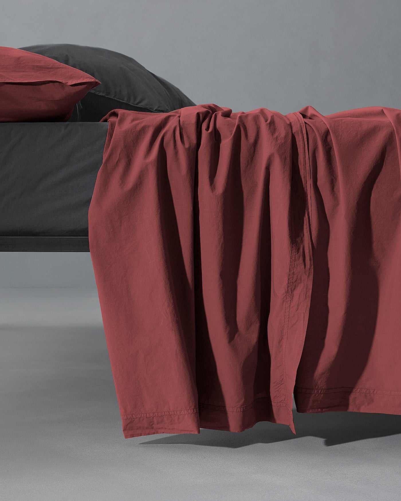 Society Limonta Nite flat sheet in terracotta, draped over bed edge against grey background.