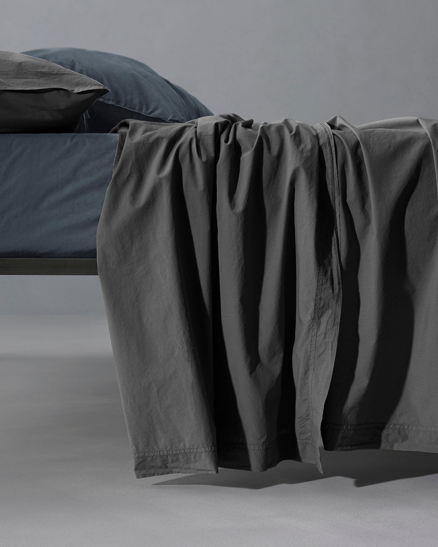 Charcoal flat sheet draped over bed edge, showing relaxed drape and subtle texture.