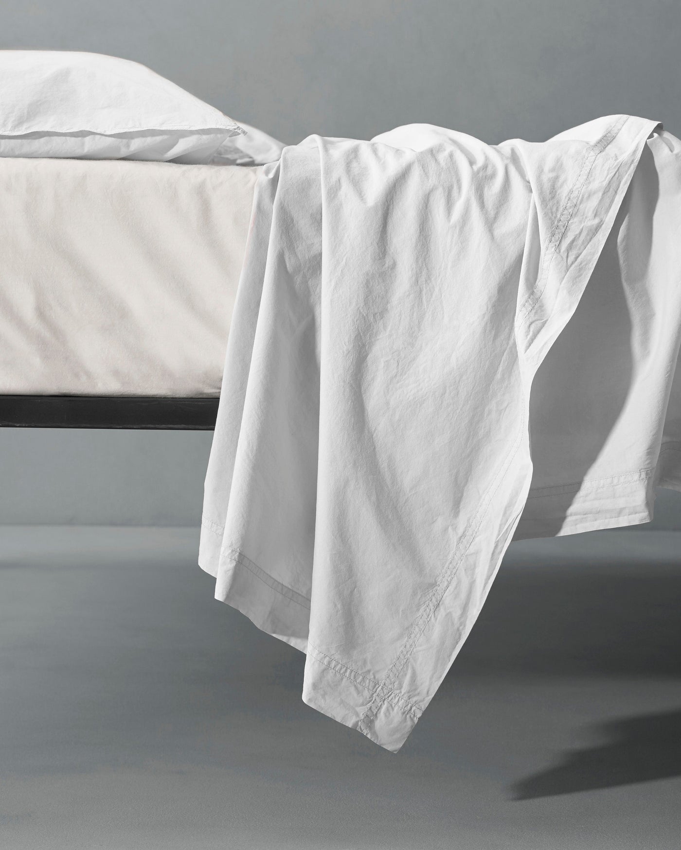 White flat sheet draped over the edge of a dark metal bed frame, pooling softly on the surface.