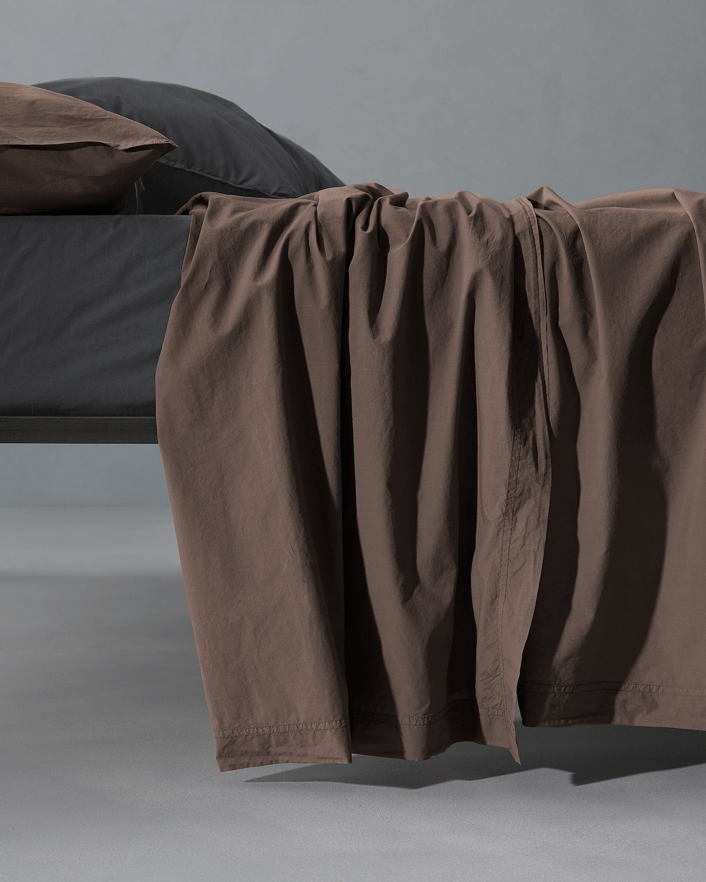 Cocoa flat sheet draped over bed edge, showing relaxed linen texture and hemstitch detail.