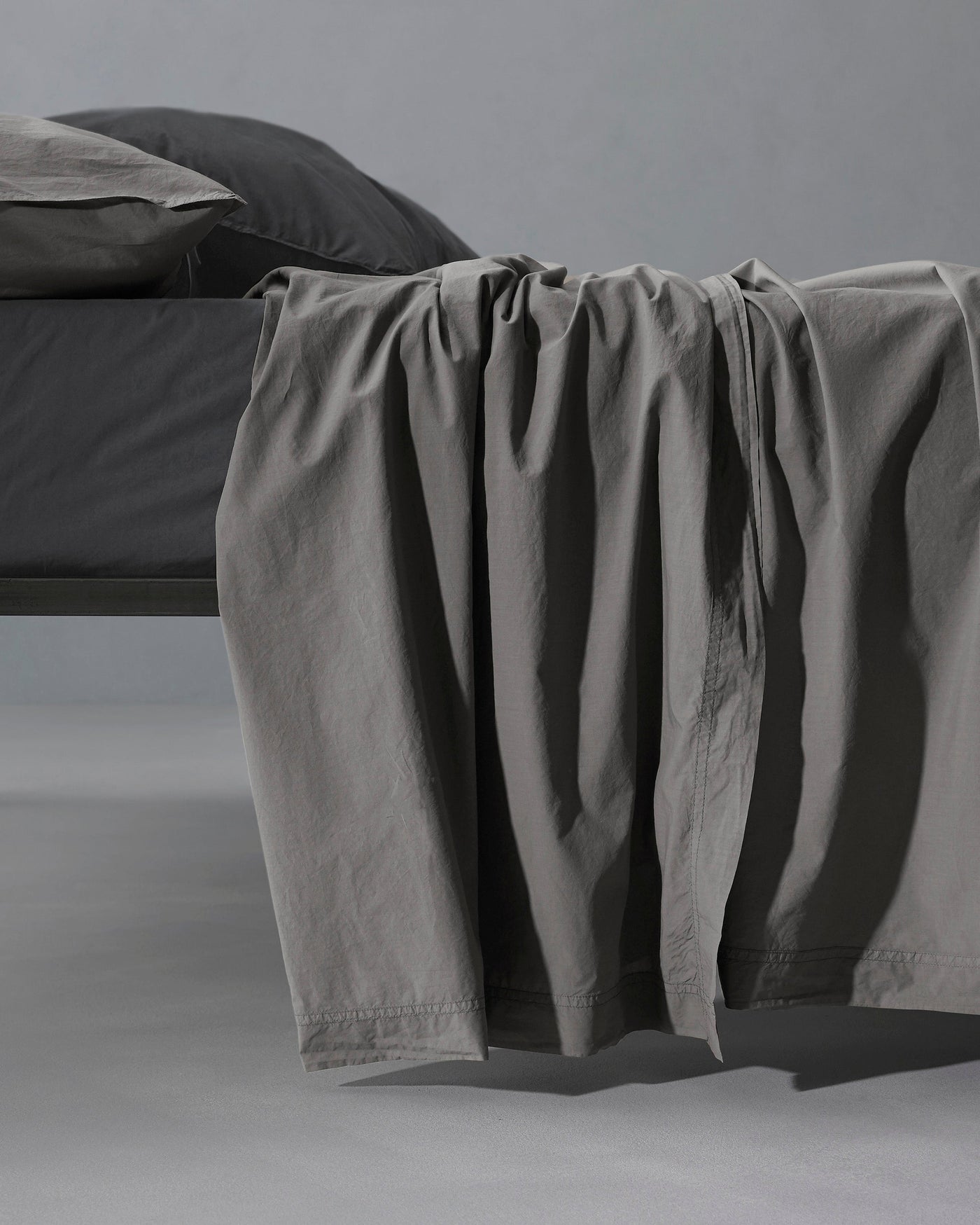 Smoke grey flat sheet draped over bed edge, showing relaxed linen texture and subtle hem detail.