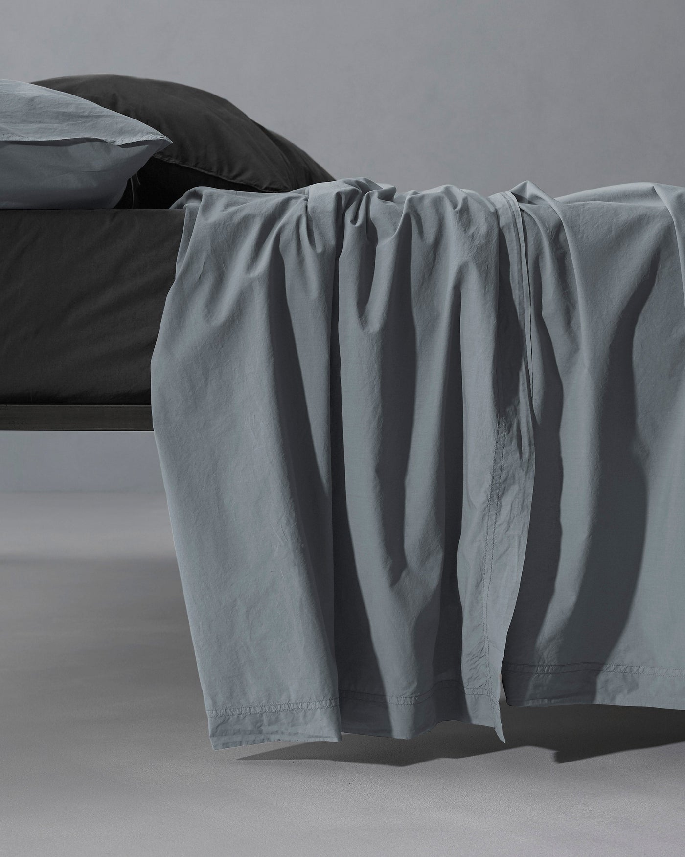 Society Limonta Nite flat sheet in slate blue draped over bed edge, showing soft drape and relaxed texture.