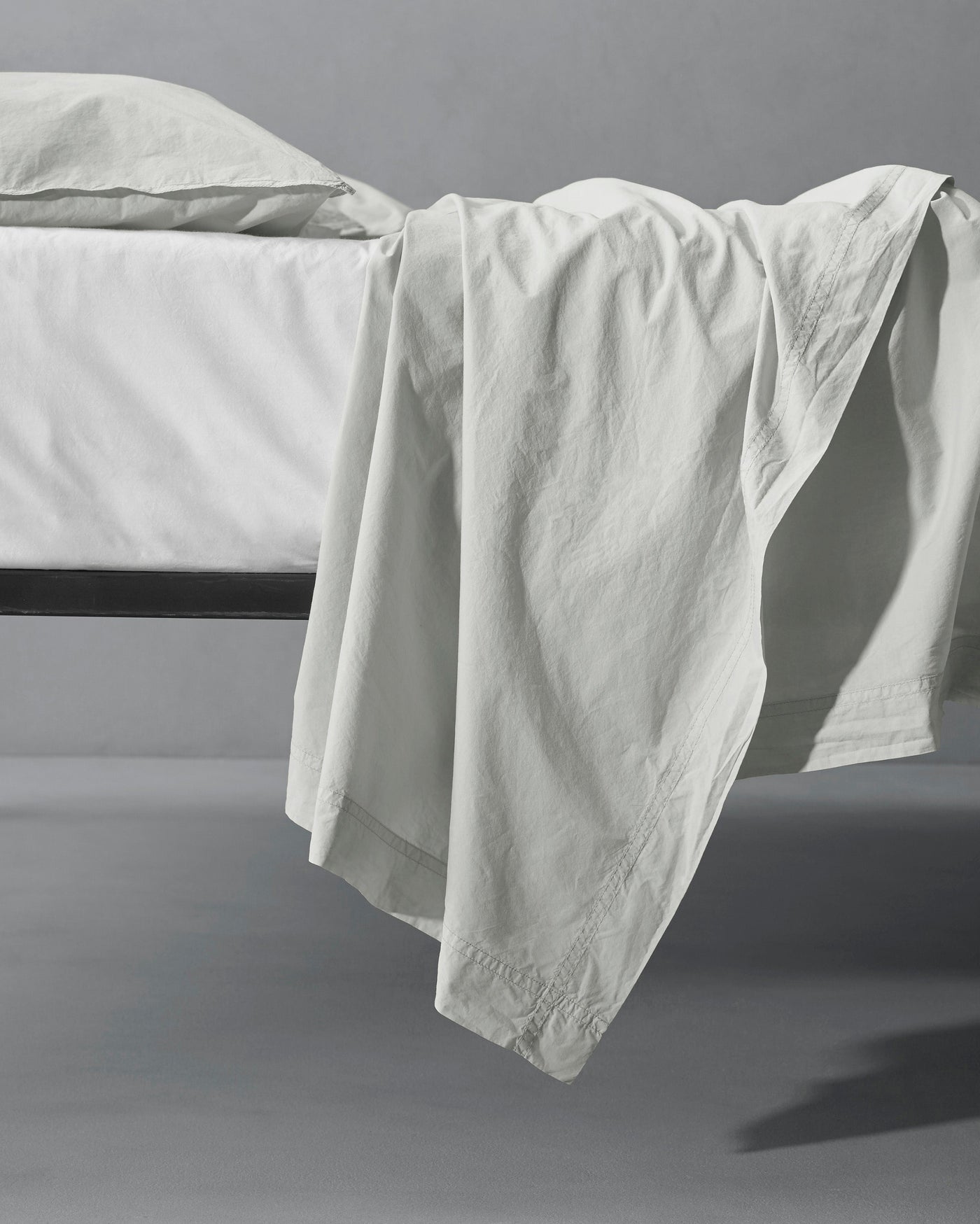 Ivory flat sheet draped over the edge of a black metal bed frame, casting soft shadows.