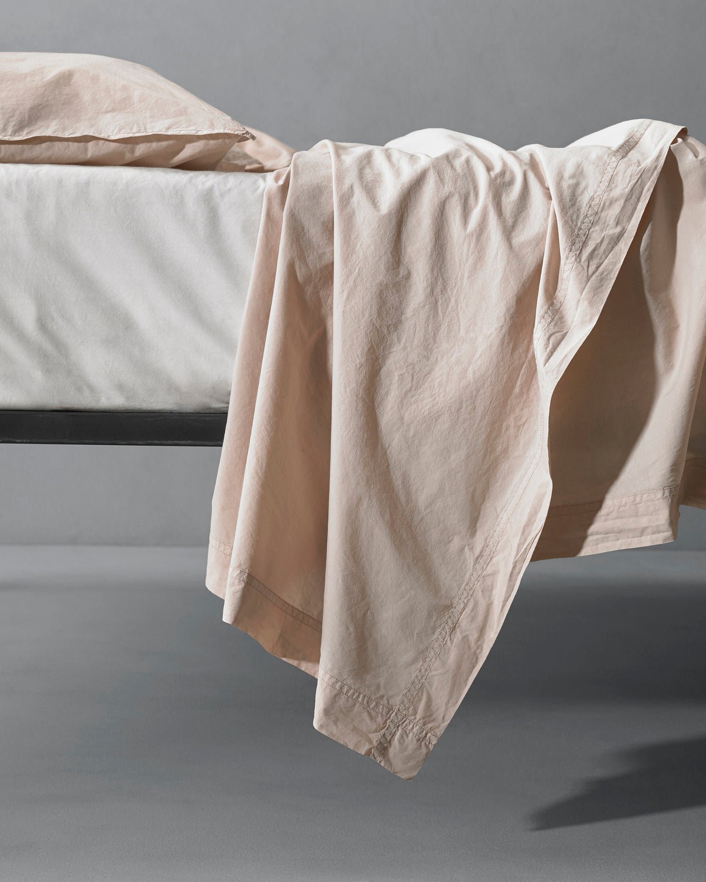 Society Limonta Nite flat sheet in blush, draped over the edge of a dark metal bed frame.