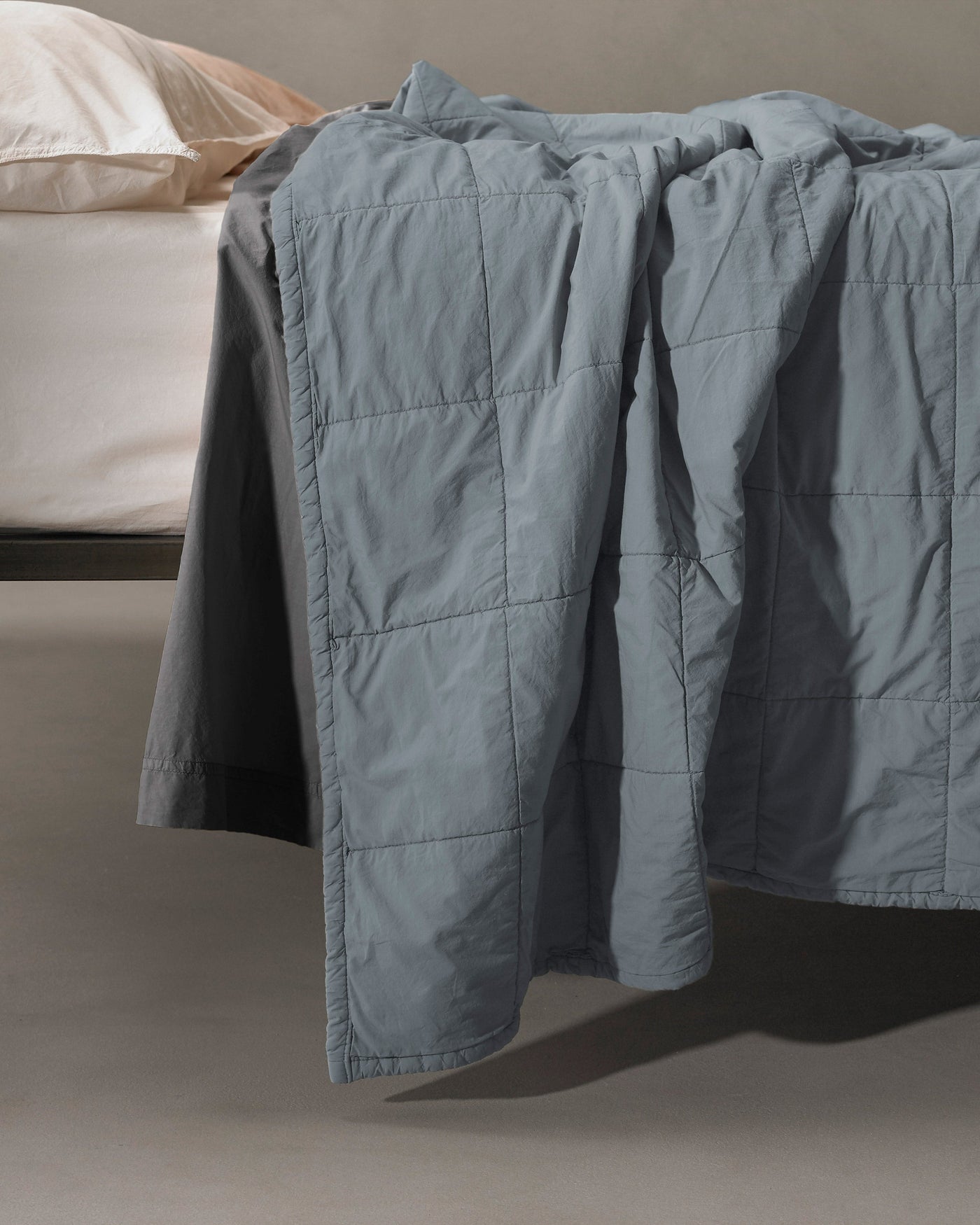 Slate blue quilted coverlet draped over bed edge, showing grid-stitch pattern and soft drape.
