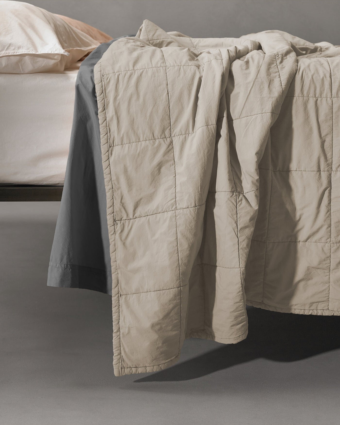 Sand-toned quilted coverlet draped over bed edge, showing grid-stitched texture and relaxed drape.