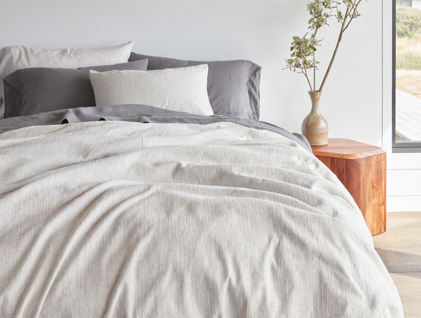 White textured duvet draped over bed with grey linen sheets, natural light setting.
