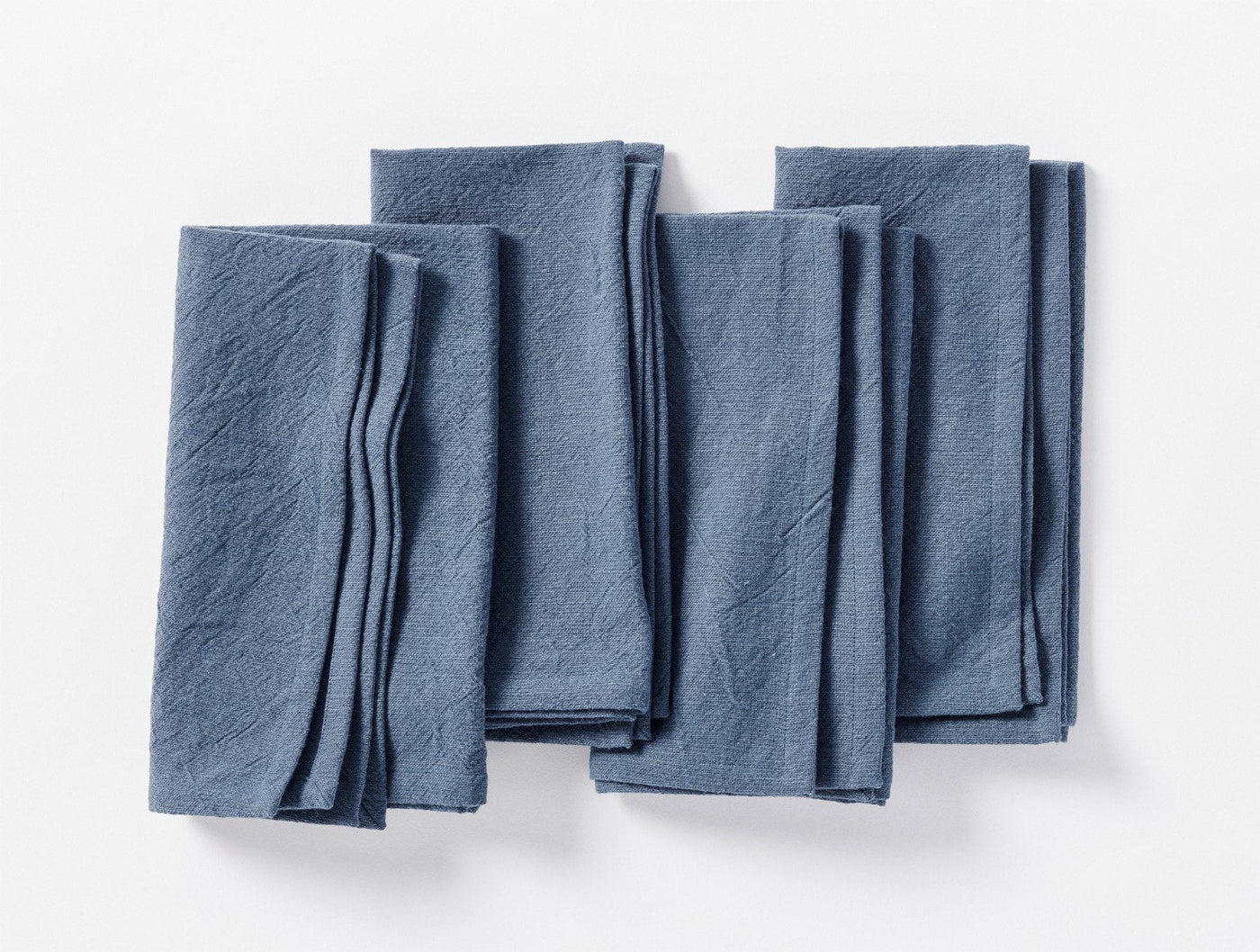Four folded Coyuchi Sonoma napkins in slate blue linen, fanned flat on white background.