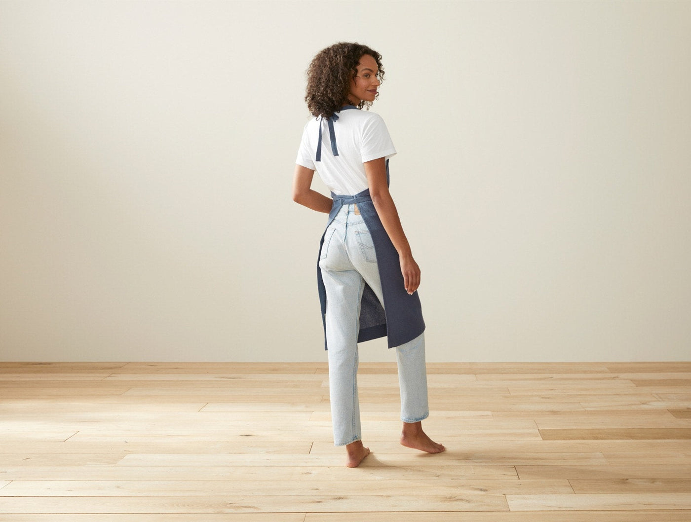 Woman modeling Coyuchi Sonoma navy blue linen apron, back view, on hardwood floor.