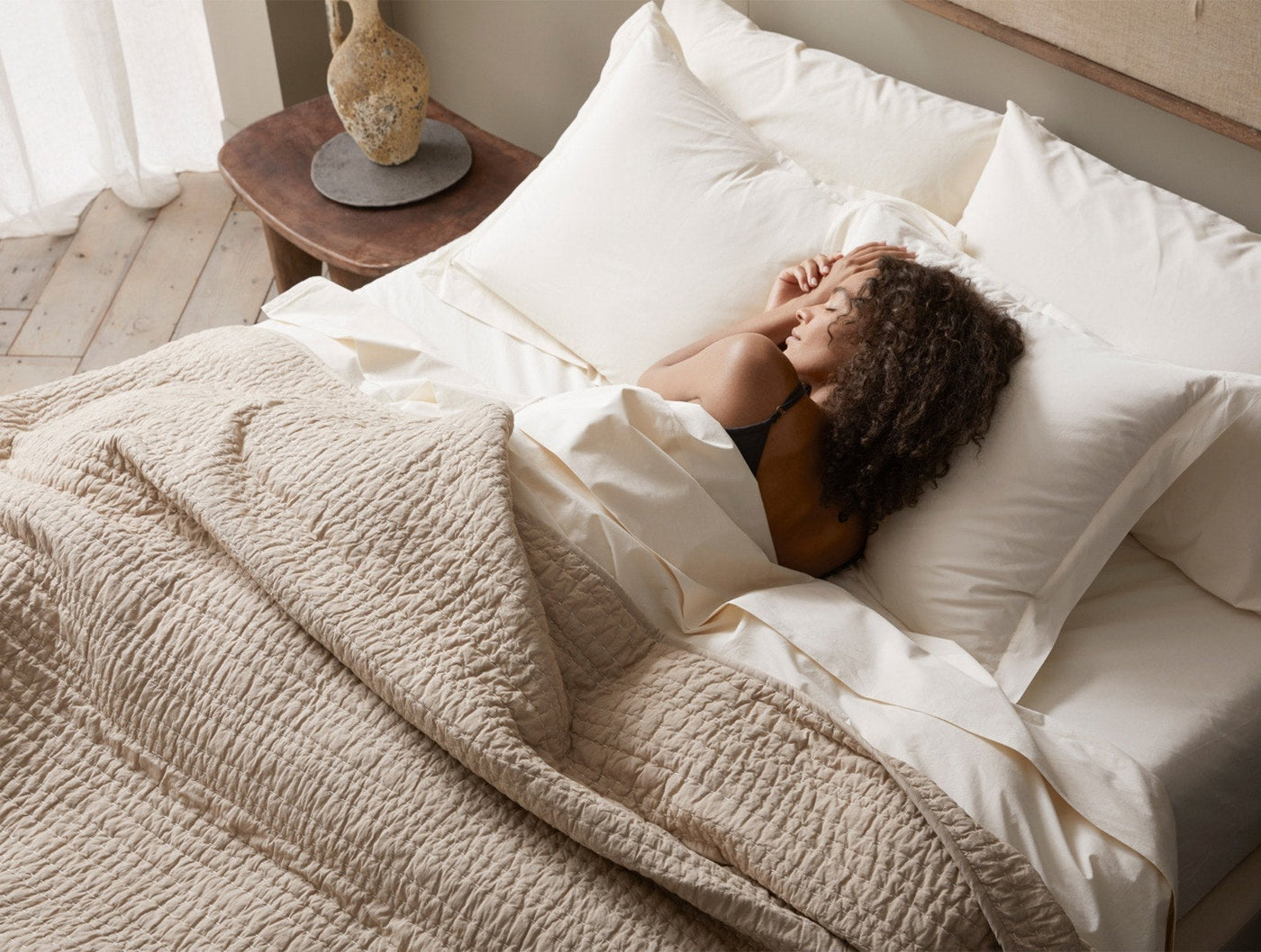 Woman resting under a Coyuchi Channel Handstitched quilt in warm linen, with ivory pillows.