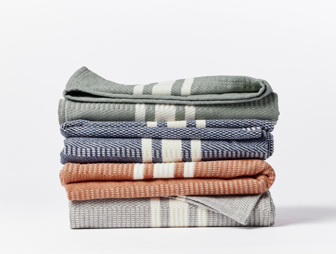 Stack of five folded Coyuchi Cirrus Supersoft throws in sage, navy, terracotta, and grey stripe.