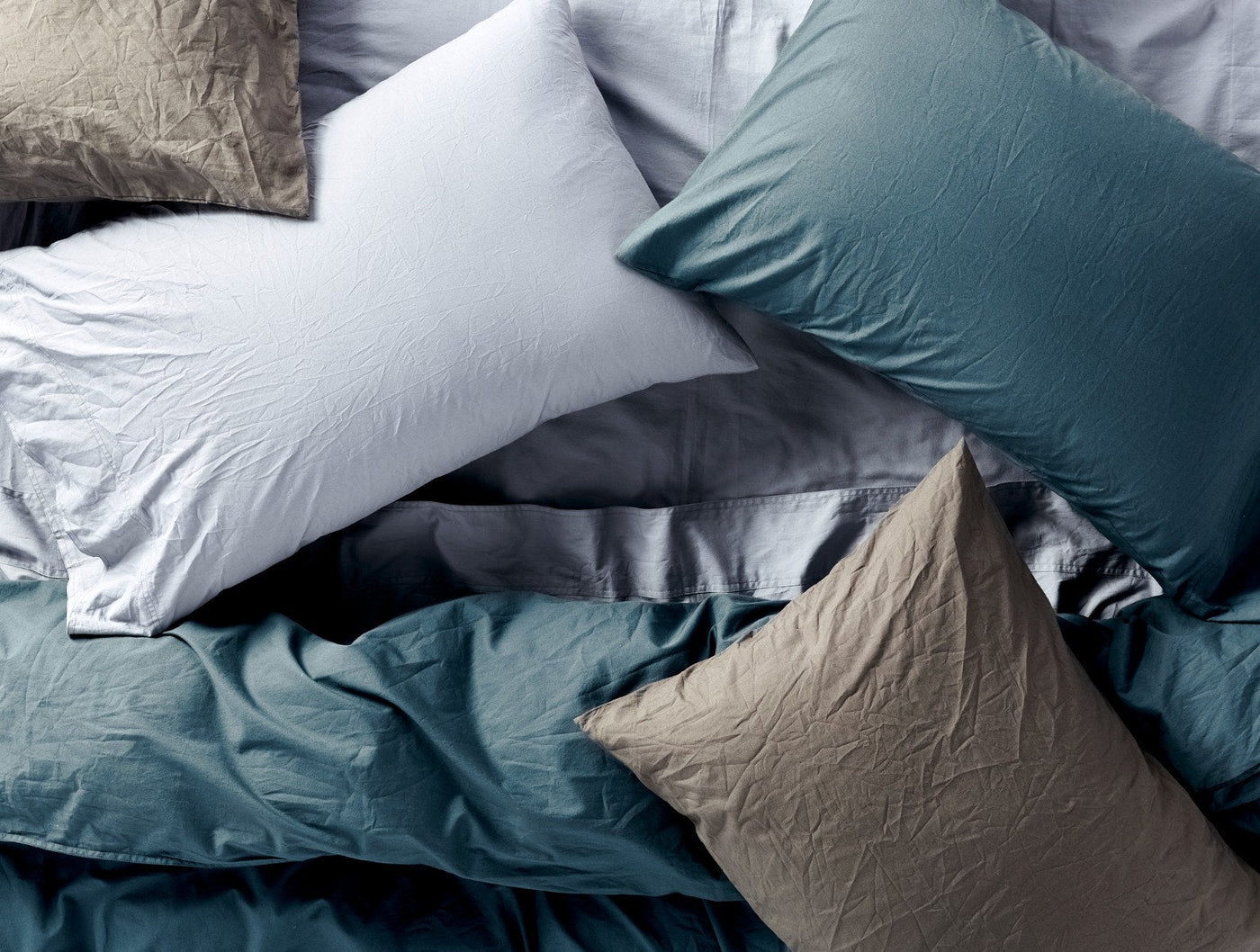 Layered crinkled percale pillows and sheets in teal, slate, and taupe tones.