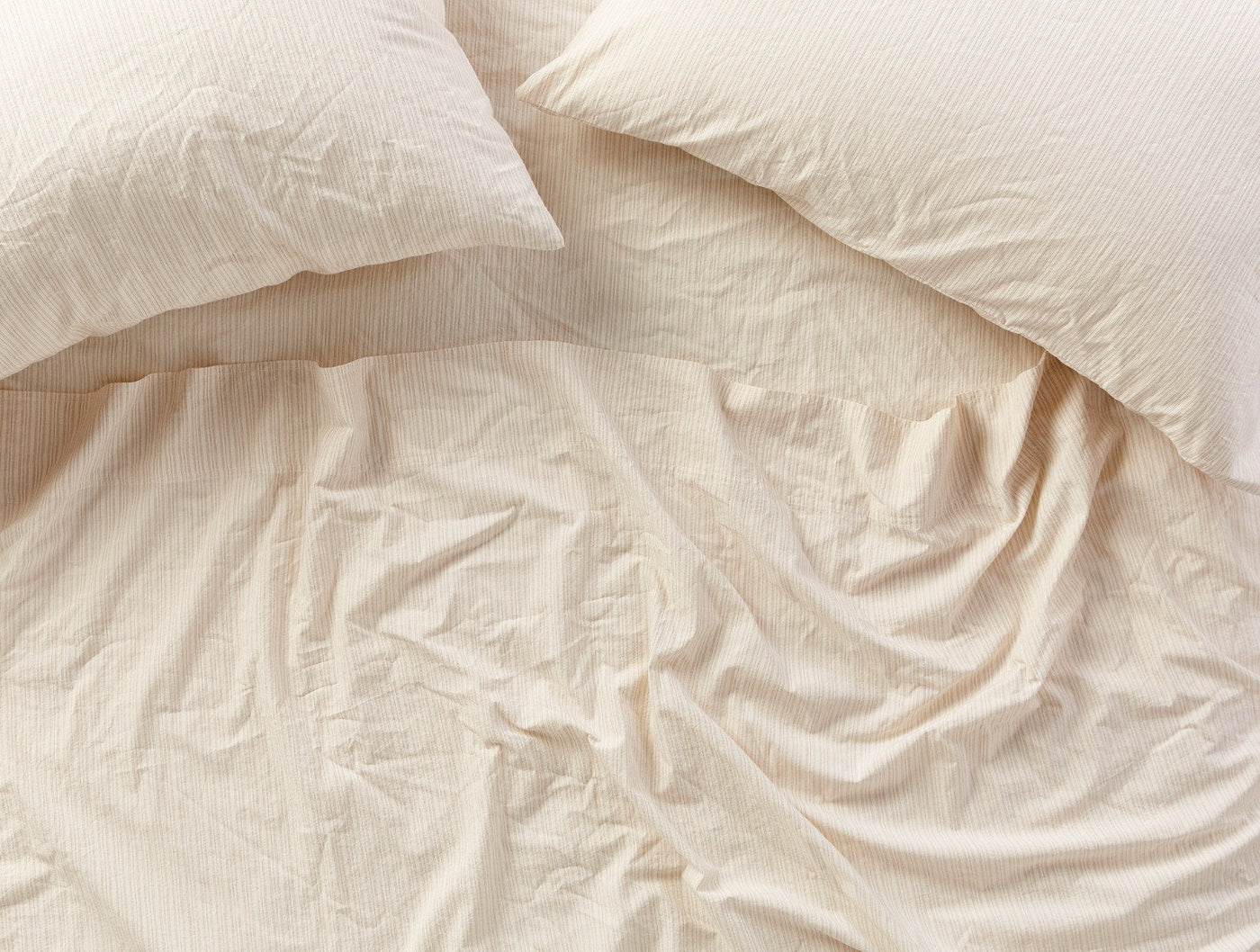 Overhead view of crinkled ivory percale fitted sheet with two pillows on a bed.
