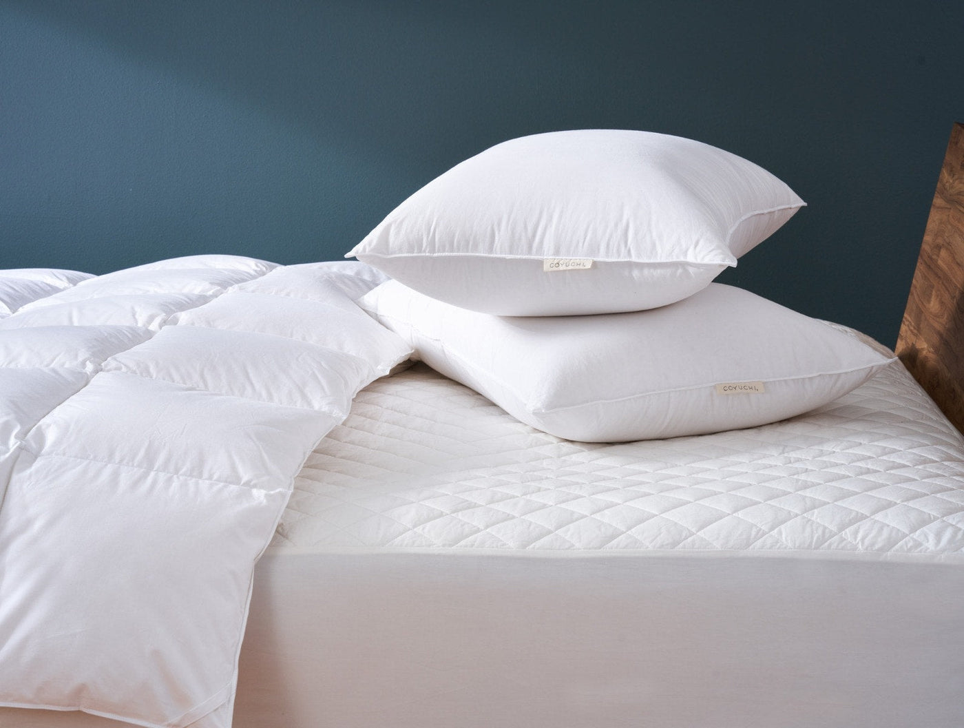 White quilted mattress pad on bed with white duvet and pillows, teal wall backdrop.