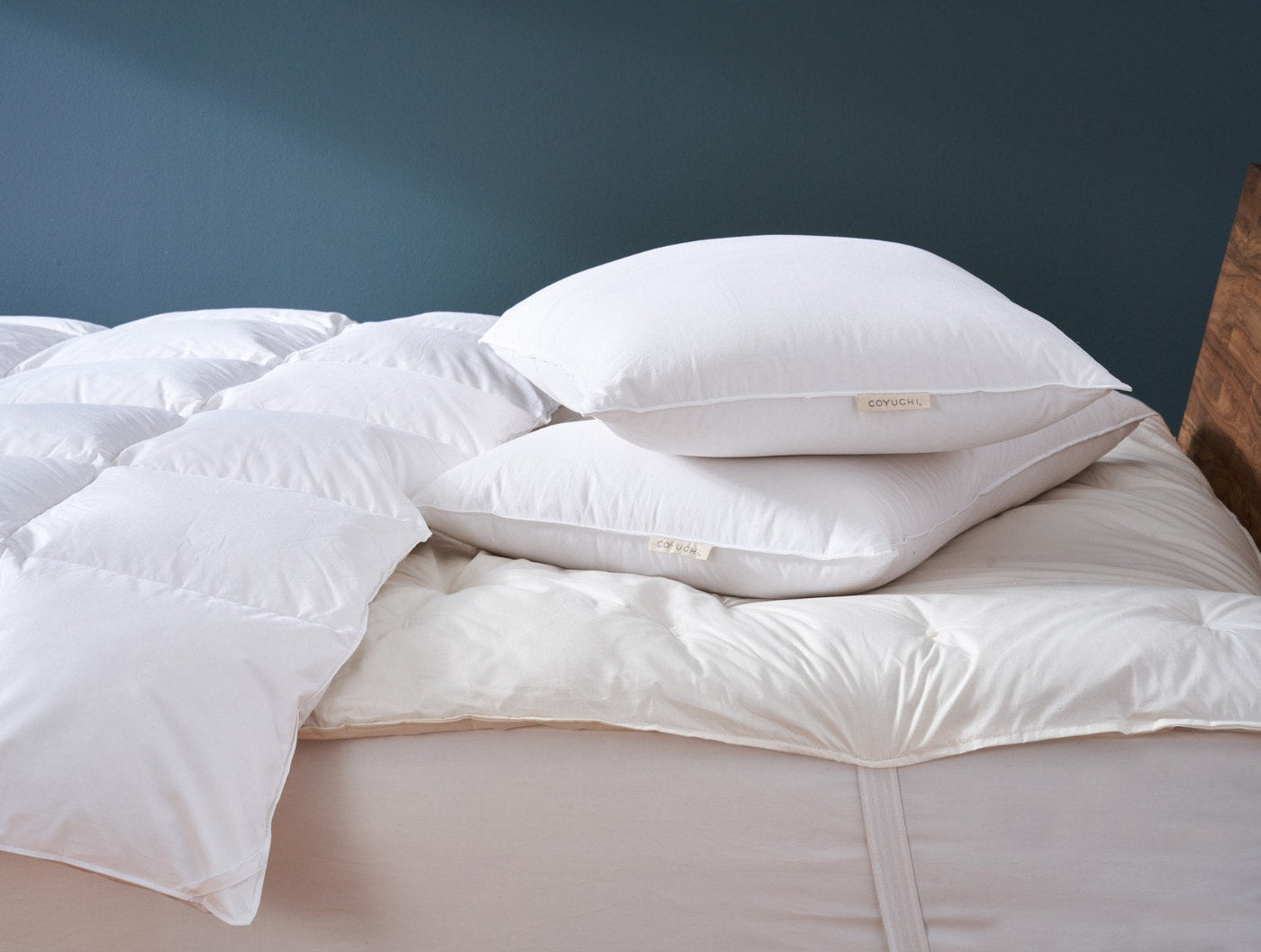 White pillows stacked on a fluffy white duvet atop a bed, teal wall backdrop.