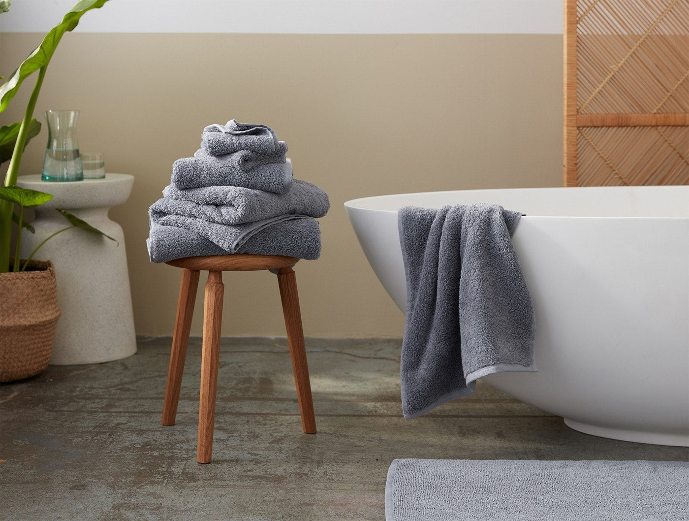 Slate grey towels stacked on wooden stool beside freestanding white bathtub.