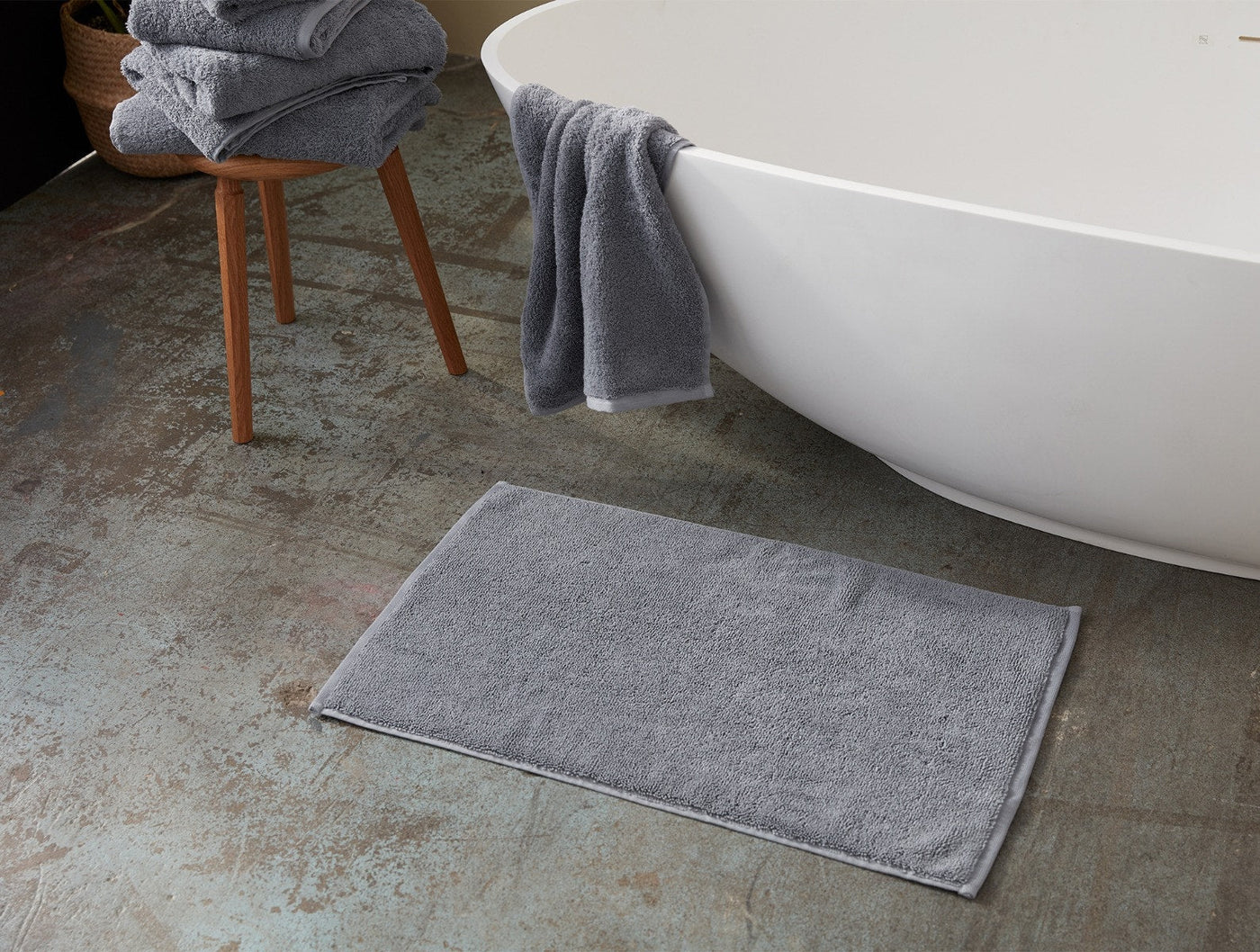 Close-up of Coyuchi Cloud Loom bath rug in grey on stone floor beside a freestanding tub.