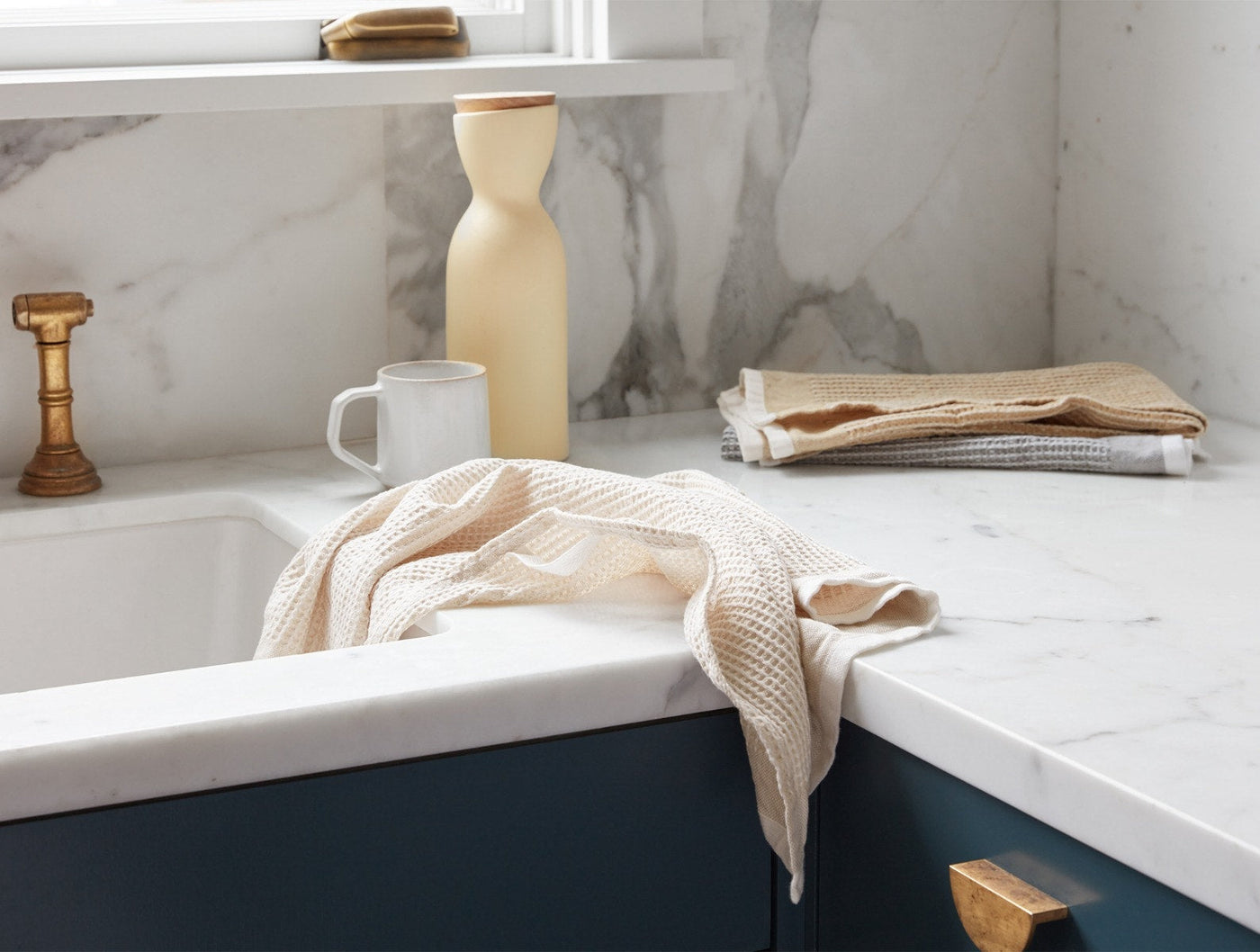 Ivory waffle-weave kitchen towel draped over navy cabinet edge, folded towels stacked on marble counter.