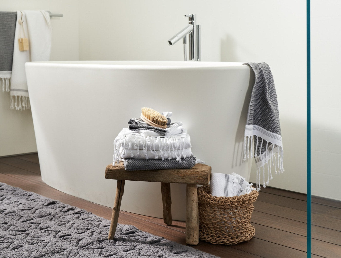 Grey and white fringed towels stacked on wooden stool beside freestanding bath.