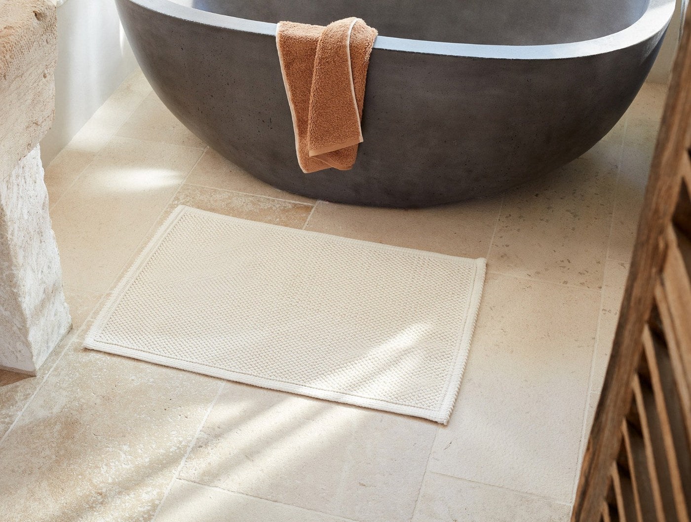 Coyuchi Pebbled bath rug in ivory, flat lay on stone floor beside a freestanding tub.