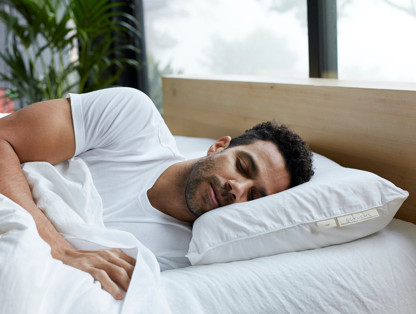Man sleeping on white Coyuchi Turiya Latex pillow, bedroom with wood headboard and plants.