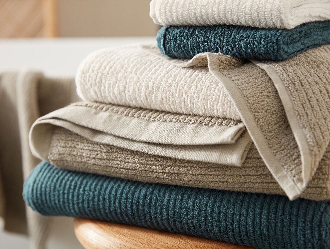 Stack of folded Coyuchi Temescal Ribbed bath towels in teal and natural tones on a wooden surface.